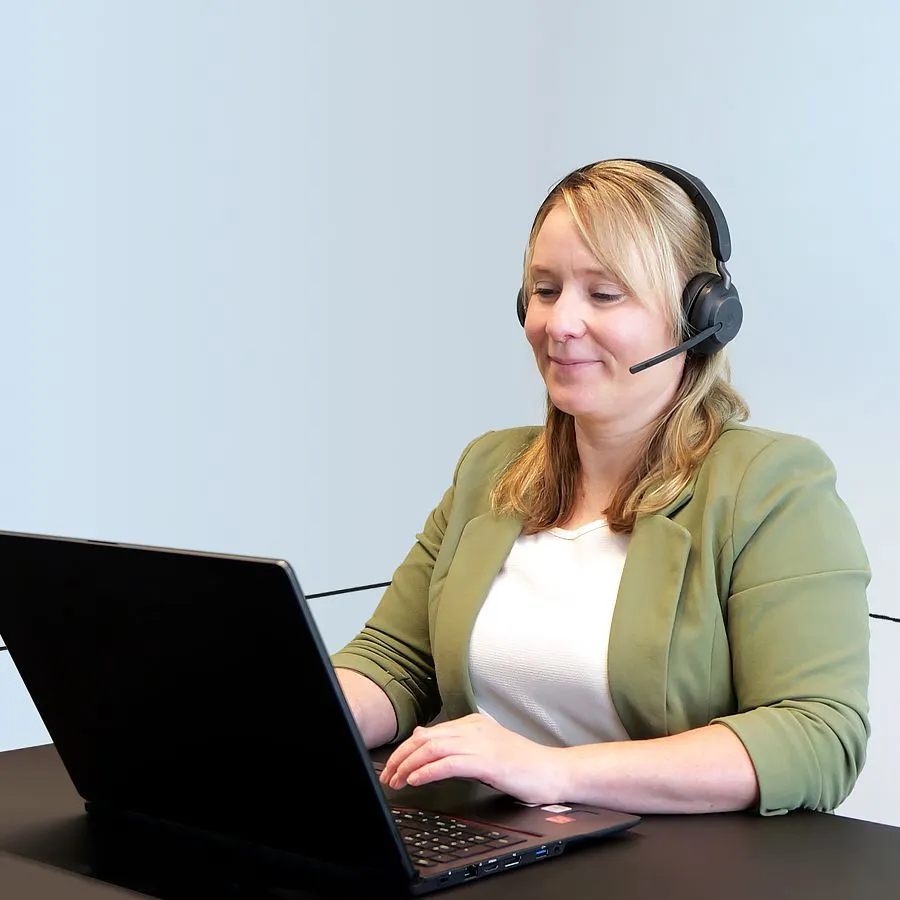 Eine lächelnde Frau mit Headset auf am Schreibtisch sitzend und am Laptop arbeitend